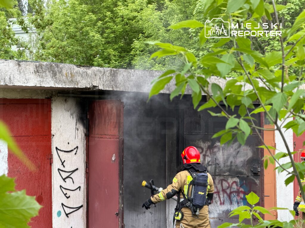 Strażak w hełmie i specjalnym ubraniu gasi dymiące zgliszcza przy czerwoną bramą budynku, otoczonego zielenią.