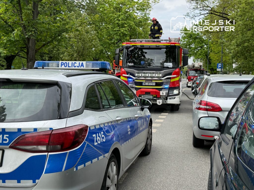 Strażak w hełmie stoi na dachu wozu strażackiego, a obok parkują radiowozy policji na zatłoczonej ulicy.