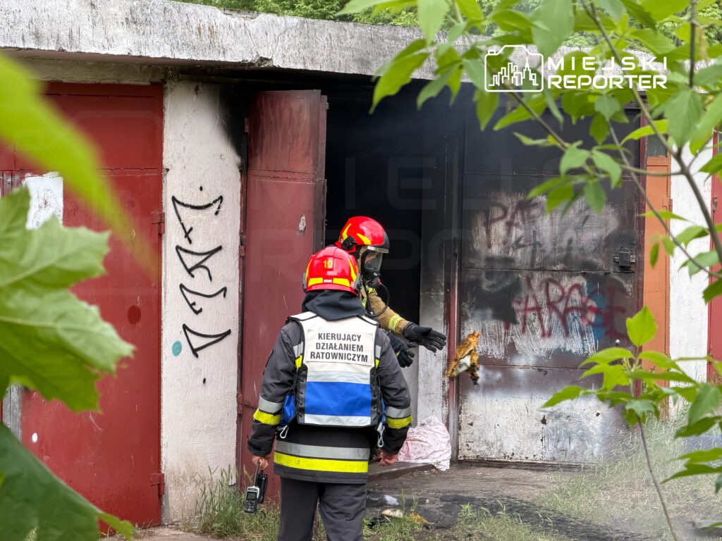 Strażacy w hełmach gaszą pożar w zniszczonym budynku, obok widoczne graffiti i dym unoszący się z wnętrza.