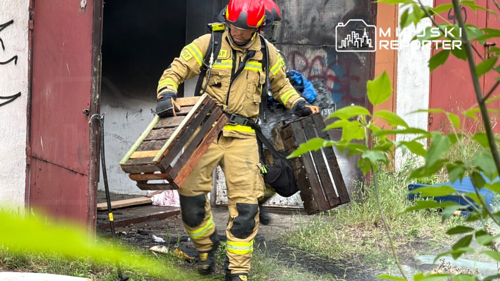 Strażak w hełmie niesie dwa drewniane palety z budynku, w którym wystąpił pożar. W tle widać zniszczone ściany.