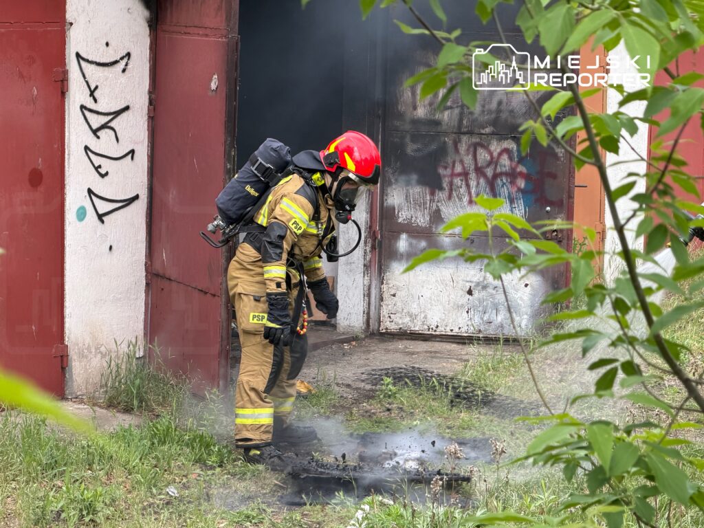 Strażak w mundurze gaśniczym gasi dymiący materiał na ziemi przed zamkniętymi drzwiami budynku.