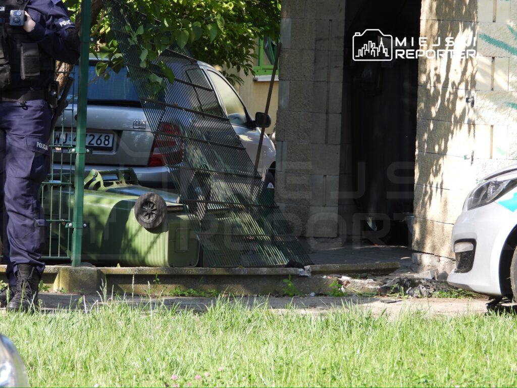 Policjant w mundurze stoi obok ogrodzenia z zielonymi pojemnikami na odpady, w tle widoczny samochód.