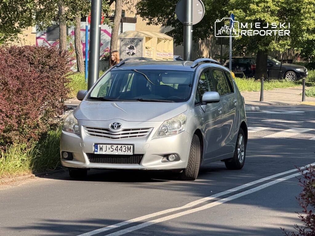 Srebrny samochód osobowy marki Toyota zaparkowany na ulicy obok zielonych krzewów i znaków drogowych.