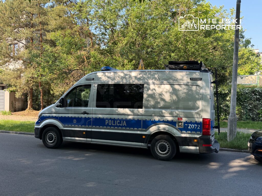 Policyjny radiowóz zaparkowany na ulicy w otoczeniu zieleni i budynków mieszkalnych.
