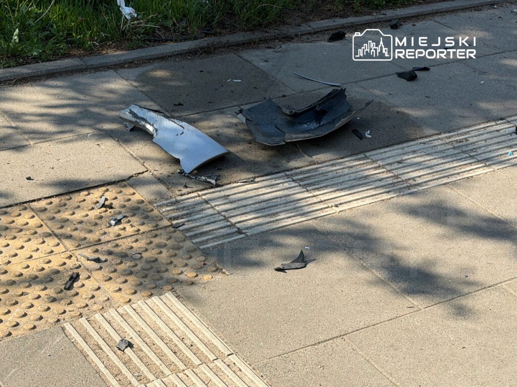 Na chodniku leżą fragmenty rozbitego pojazdu oraz drobne odpady, w tle widoczna zieleń.