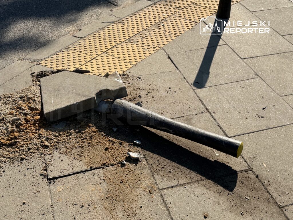 Złamany słup sygnalizacyjny leży na chodniku obok uszkodzonej płyty betonowej, wokół widać piasek i gruz.