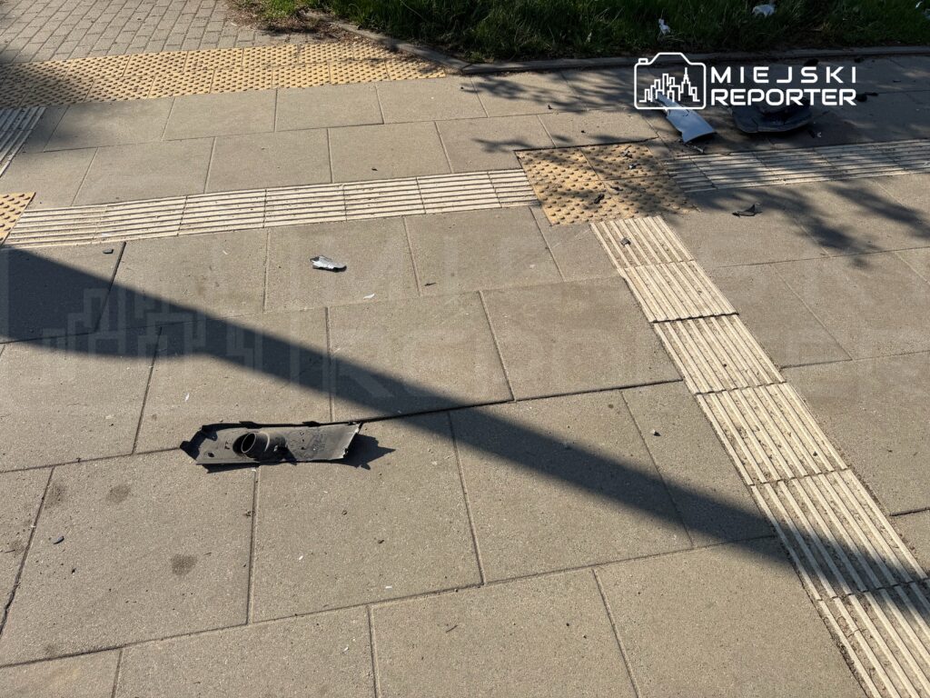 Uszkodzony fragment chodnika z metalowym elementem leżącym na płytkach, obok widać drobne śmieci.