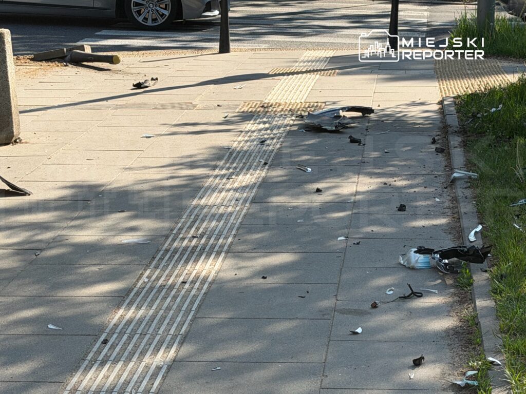 Na chodniku leżą fragmenty zniszczonego przedmiotu oraz odłamki, w tle widoczny samochód.