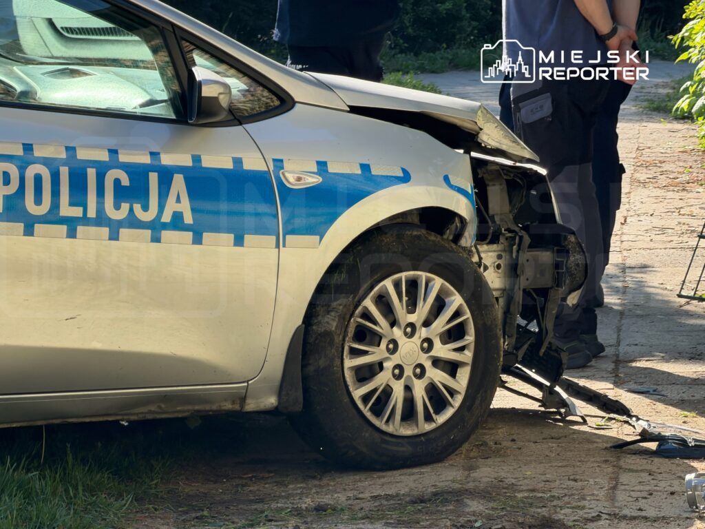 Uszkodzony radiowóz policyjny z uszkodzoną przednią częścią stoi na poboczu, obok dwóch policjantów.