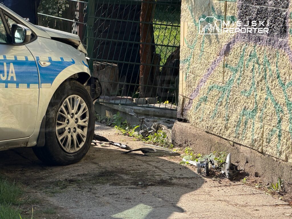 Policyjny samochód zaparkowany obok uszkodzonej ściany budynku, z widocznymi śladami graffiti i zarośniętą nawierzchnią.