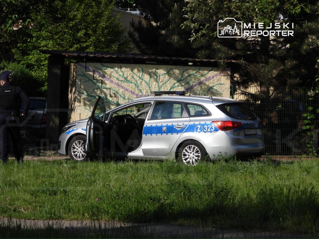 Policjant w mundurze stoi obok otwartego radiowozu z niebieskimi oznaczeniami na trawniku.