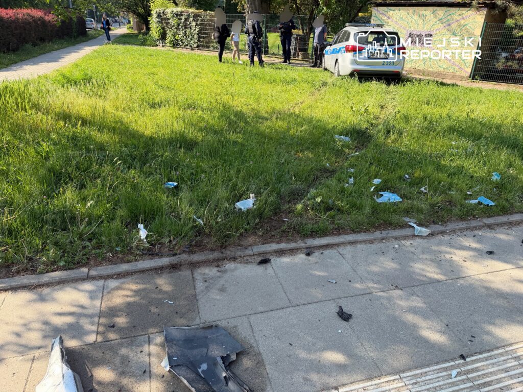 Grupa policjantów zbiera dowody na trawniku, obok rozbitego pojazdu i porozrzucanych fragmentów.