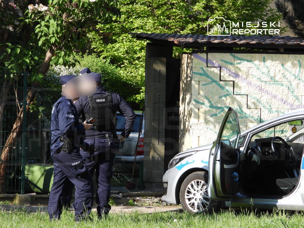 Dwóch policjantów w mundurach rozmawia obok otwartego samochodu w zielonym otoczeniu.