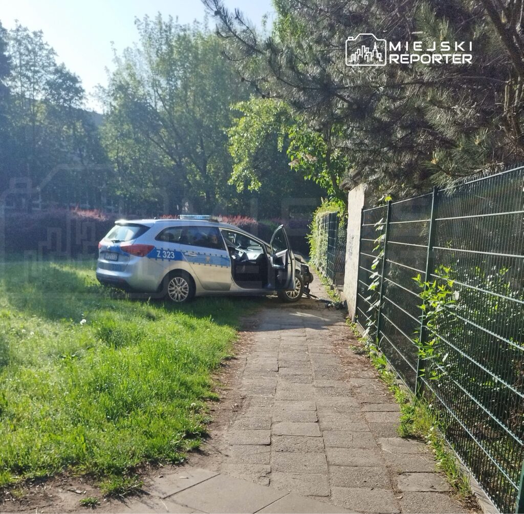 Policyjny radiowóz zaparkowany na trawniku z otwartymi drzwiami obok ogrodzenia w słonecznym otoczeniu.