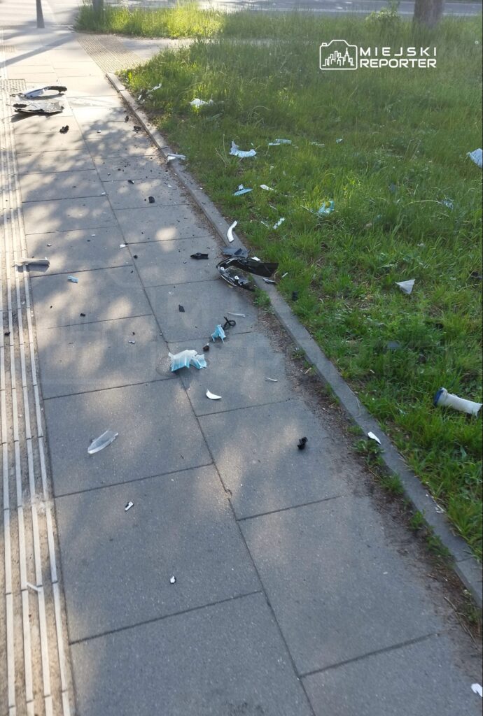 Na chodniku leżą porozrzucane odpady, w tym fragmenty plastików i papierów, obok trawnika.