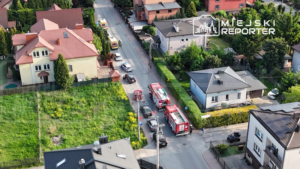 Na ulicy stoją dwa wozy strażackie, a obok nich policja i kilka samochodów cywilnych. W tle widać domy jednorodzinne.