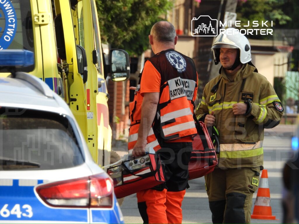 Ratownicy medyczni rozmawiają z strażakiem na ulicy, obok pojazdów służb ratunkowych.