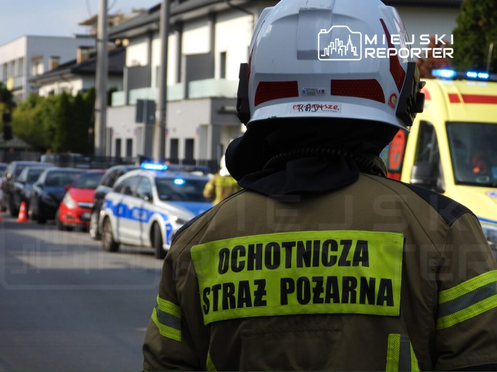Strażak w mundurze z napisem "OCHOTNICZA STRAŻ POŻARNA" obserwuje akcję ratunkową w mieście, w tle radiowóz policji.