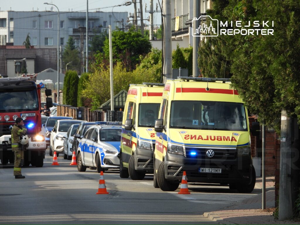Strażak w mundurze rozmawia przez radio obok ambulansów i radiowozu policyjnego na zatłoczonej ulicy.