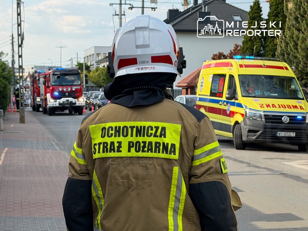Strażak w uniformie obserwujący akcję ratunkową na ulicy, obok przejeżdżające pojazdy straży pożarnej i karetka.
