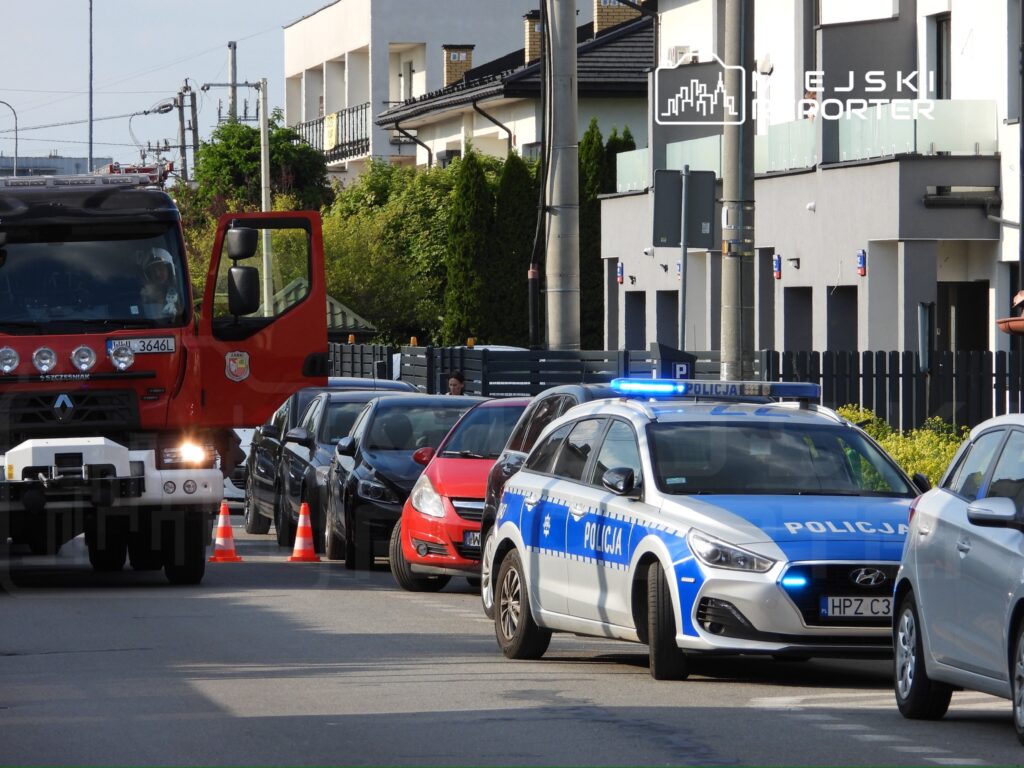 Wozidło strażackie z otwartymi drzwiami stoi na ulicy, obok policyjny radiowóz z włączonymi sygnałami świetlnymi.