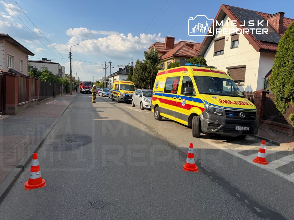 Na ulicy parkują karetka pogotowia, woz strażacki i radiowóz, a obok stoi strażak kierujący ruchem.