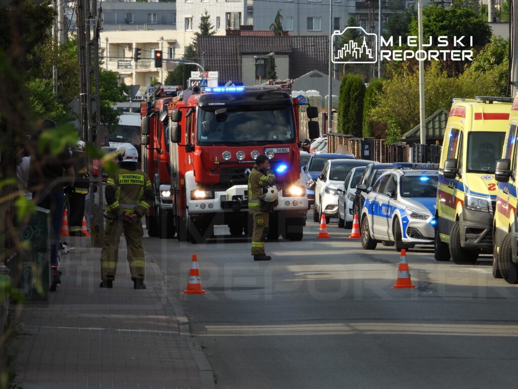 Strażacy w mundurach i kaskach stoją obok wozu strażackiego, a w tle widać policyjny radiowóz oraz karetki.