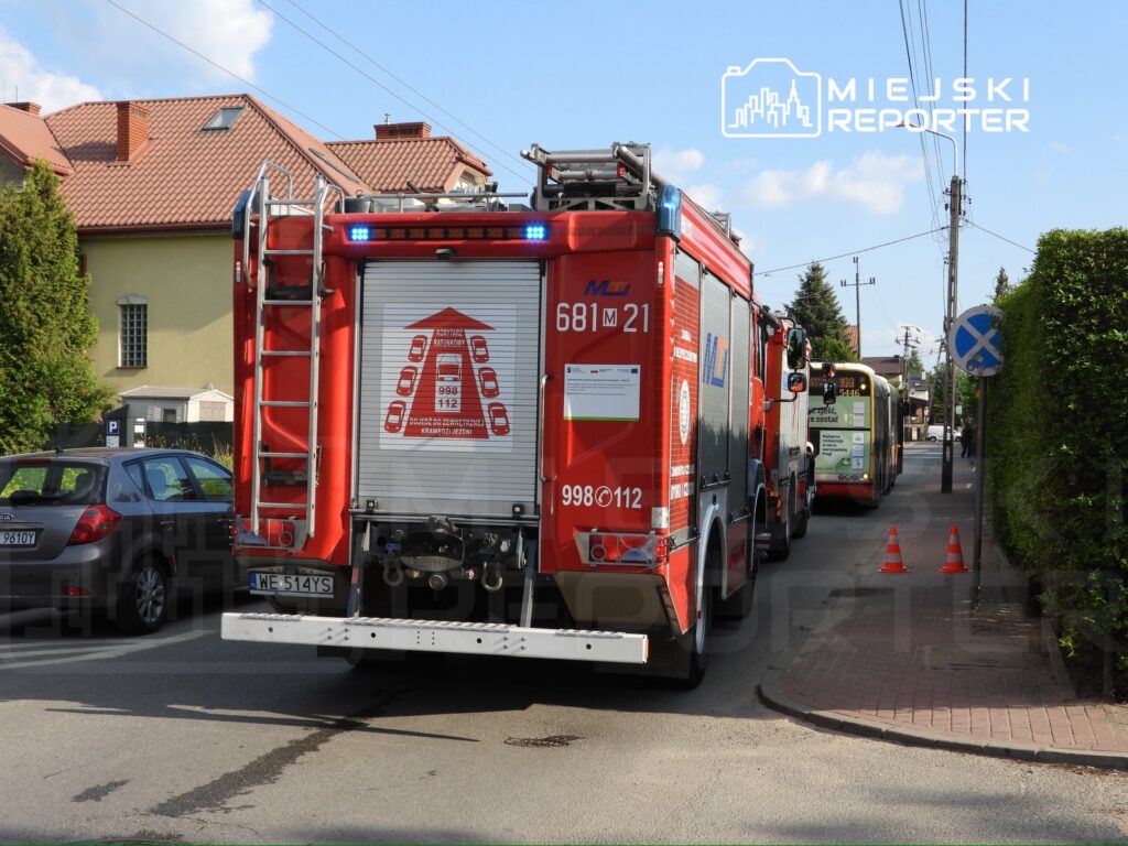 Czerwony wóz strażacki z włączonymi sygnalizatorami stoi na ulicy, obok zaparkowane samochody i autobus.