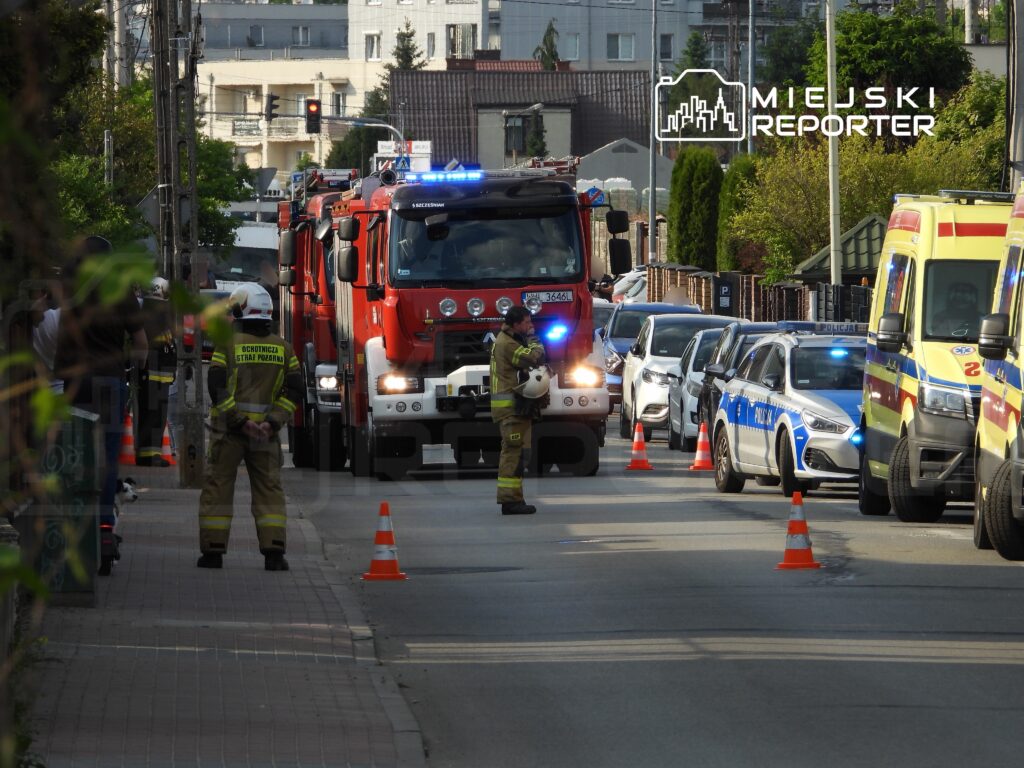 Strażacy w mundurach kontrolują ruch przy zablokowanej ulicy, obok pojazdy straży pożarnej i policji.