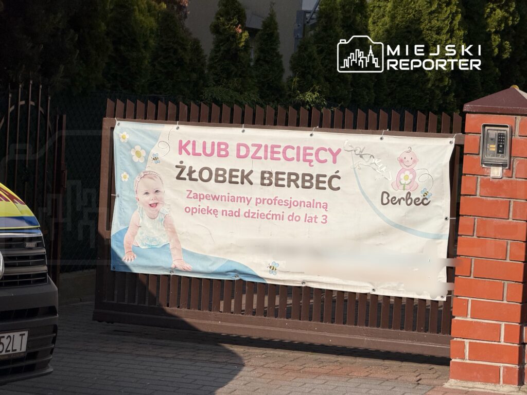 Baner informacyjny z napisem "KLUB DZIECIĘCY ŻŁOBEK BERBEC" oraz hasłem "Zapewniamy profesjonalną opiekę nad dziećmi do lat 3" umieszczony na ogrodzeniu.