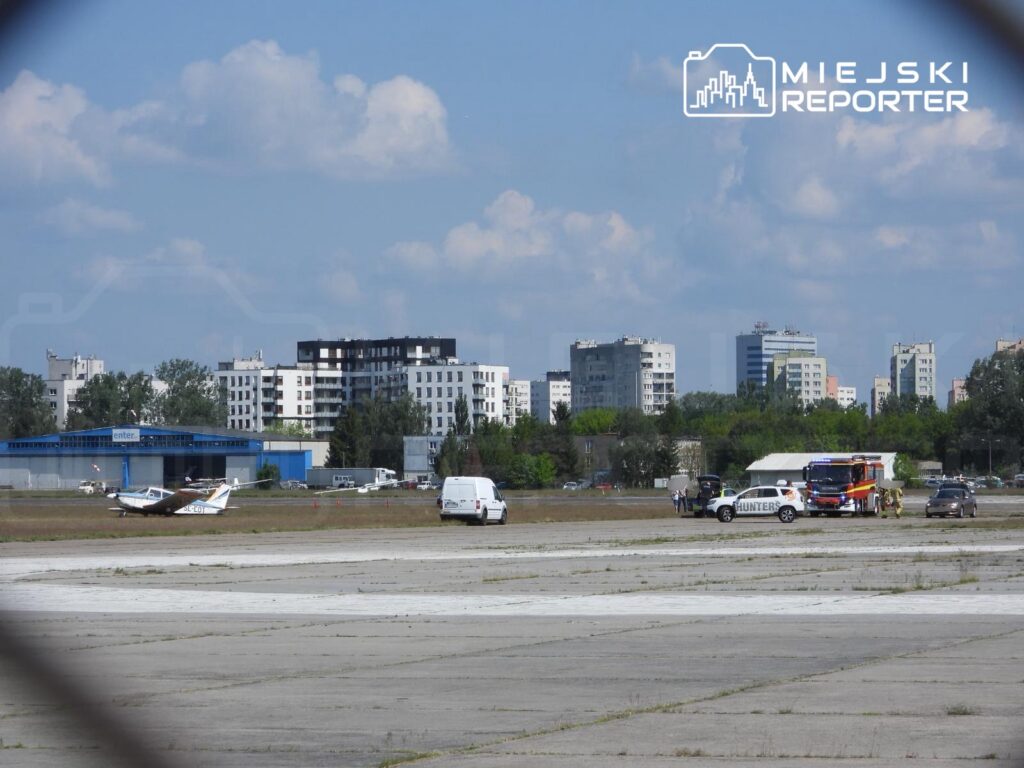 Mały samolot stoi na pasie startowym, obok zespół pojazdów ratunkowych i kilka osób w tle. W oddali nowoczesne budynki.