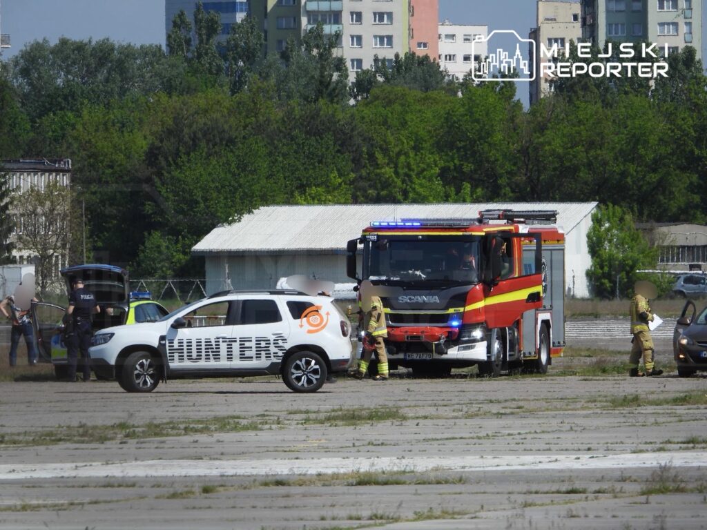 Na terenie byłego lotniska, strażacy i policjanci interweniują przy zaparkowanych samochodach, w tym radiowozie.