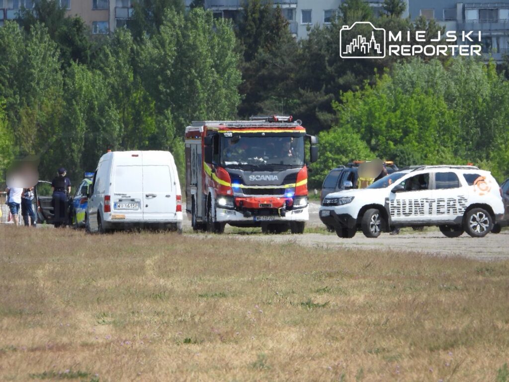 Grupa osób rozmawia z policjantami obok białego furgonu i pojazdu straży pożarnej na terenie zielonym.