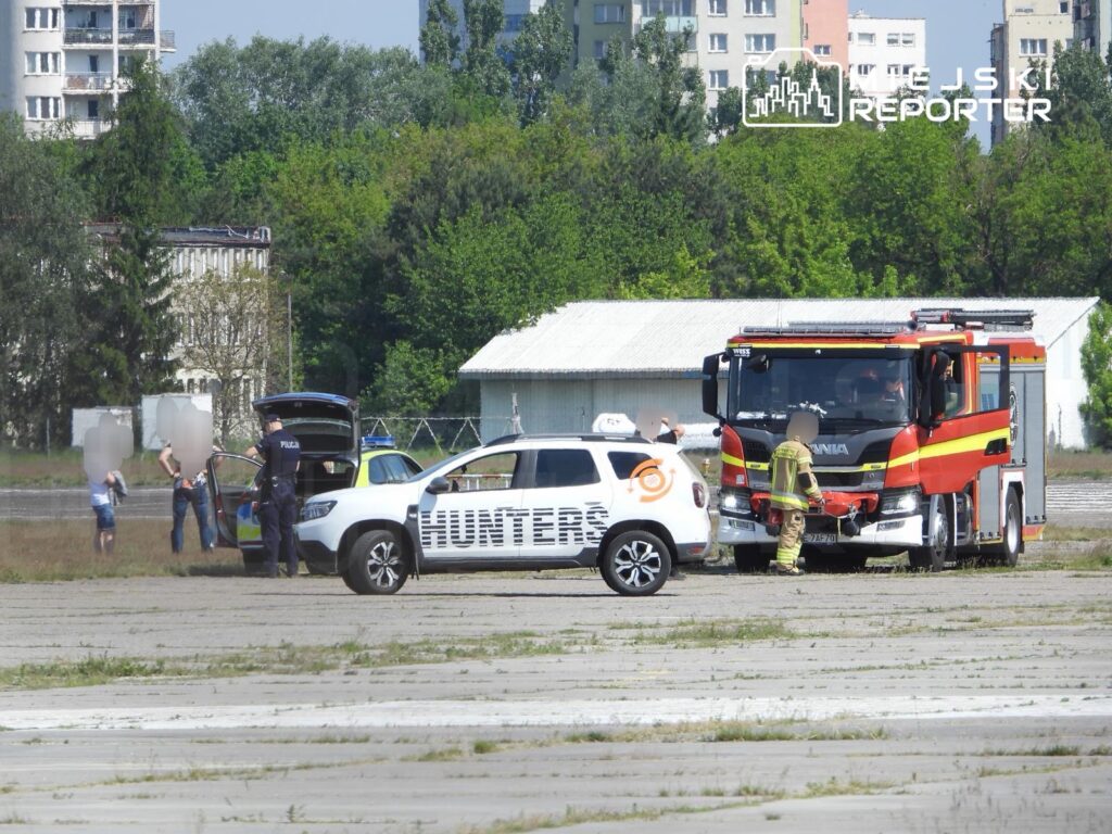 Policjant rozmawia z dwoma osobami obok radiowozu, podczas gdy strażak sprawdza wóz strażacki na opuszczonym lotnisku.