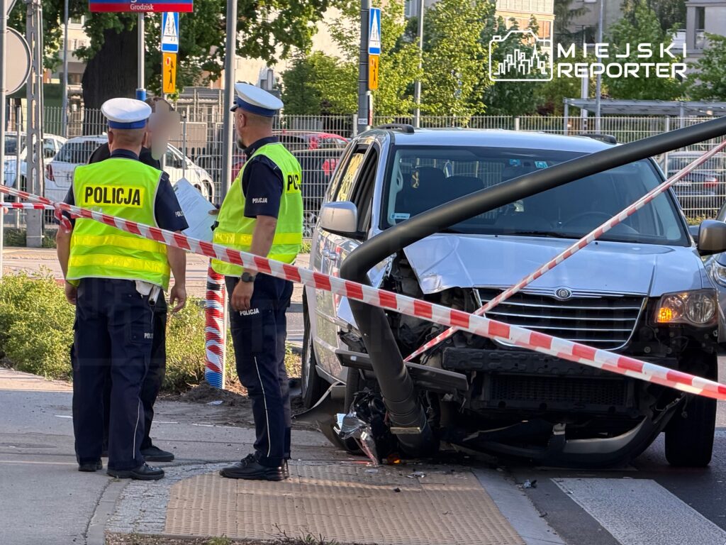 Policjanci w kamizelkach odblaskowych prowadzą czynności na miejscu wypadku drogowego z uszkodzonym samochodem i latarnią.