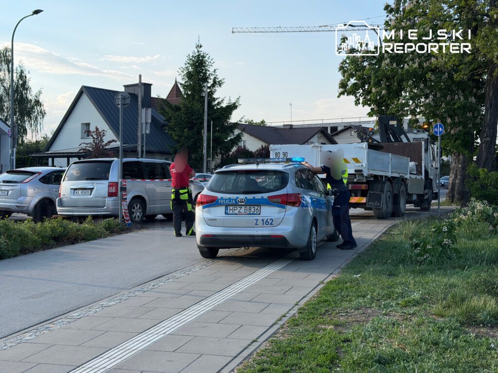 Policjant rozmawia z kierowcą przy radiowozie na ulicy, obok parkujących samochodów i ciężarówki.