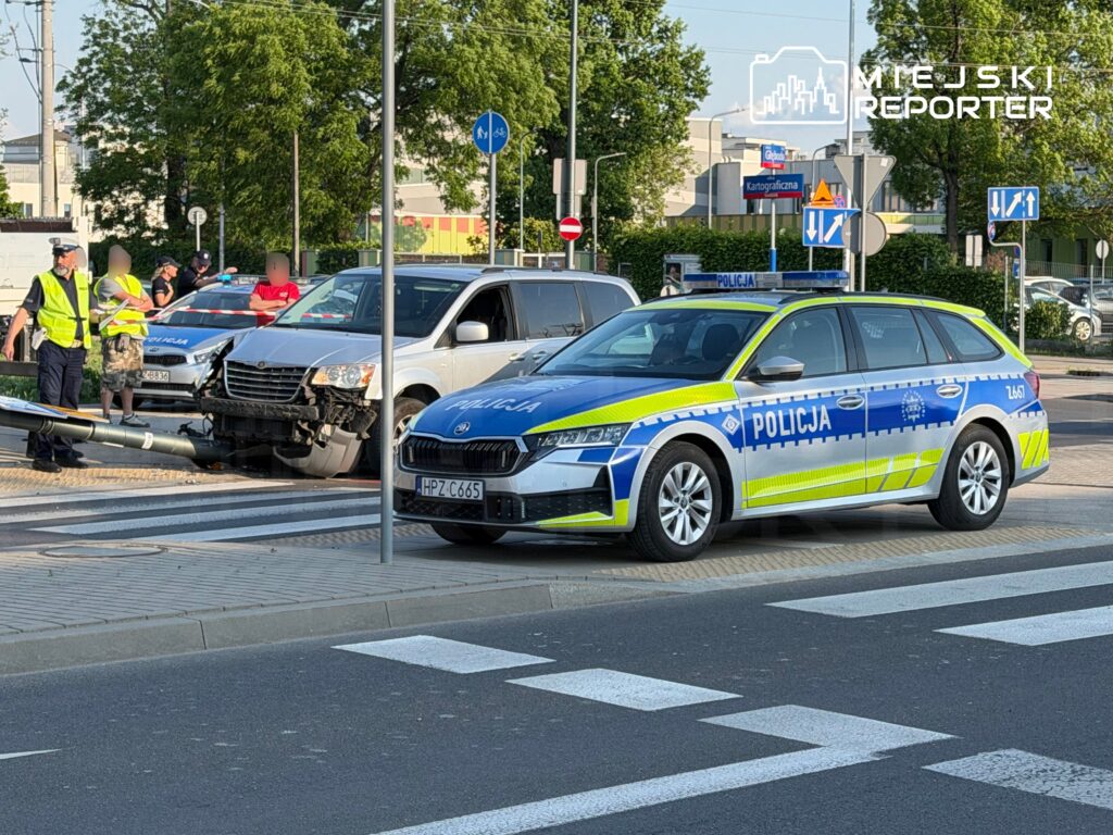 Na skrzyżowaniu stoi radiowóz policyjny, a obok grupka osób oraz uszkodzony samochód z rozbitym przodem.