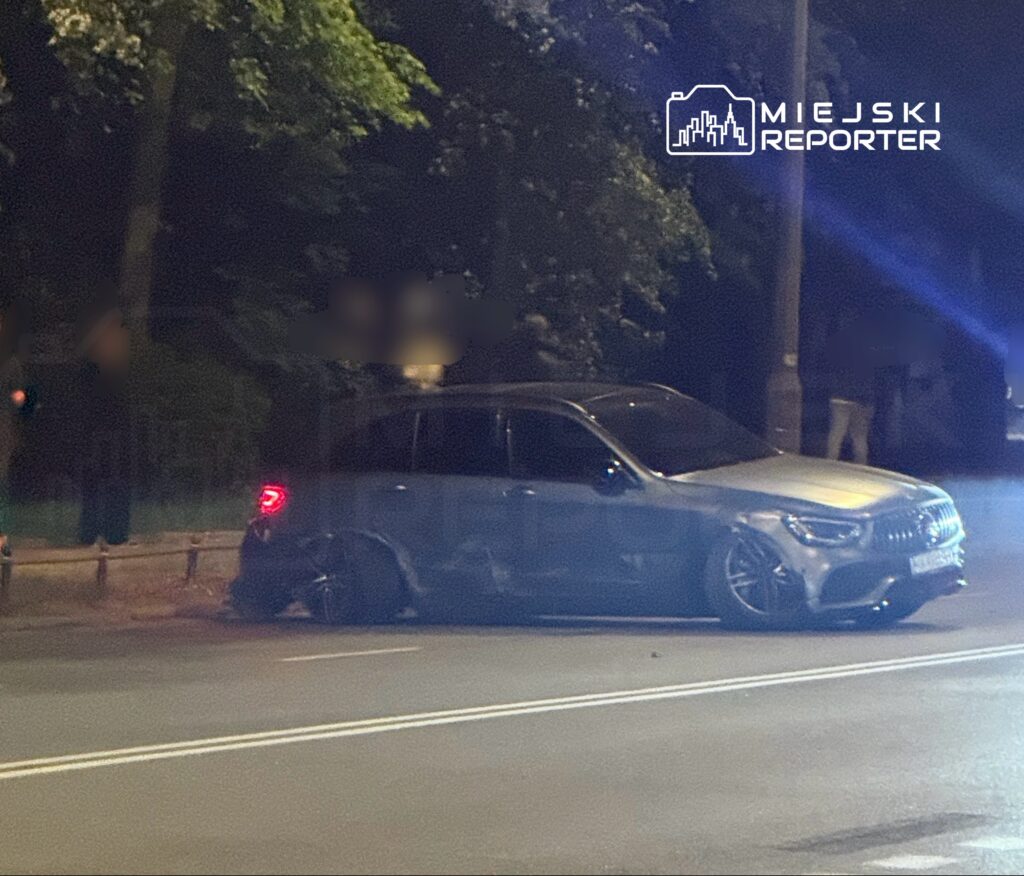 Uszkodzony samochód osobowy zaparkowany na poboczu ulicy, obok grupy osób w ciemnym otoczeniu.