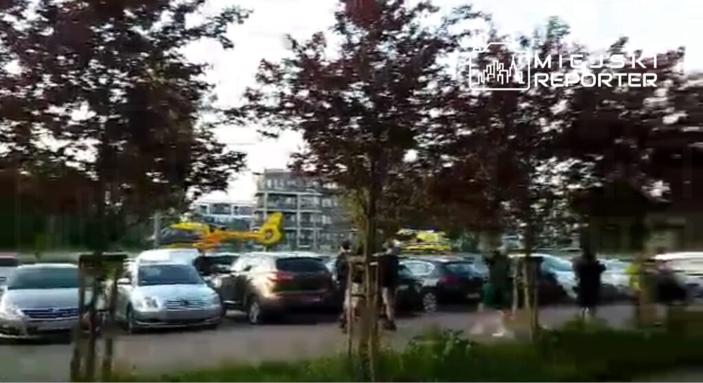 Helikopter ratunkowy ląduje na parkingu otoczonym samochodami i drzewami, w tle widoczny nowoczesny budynek.