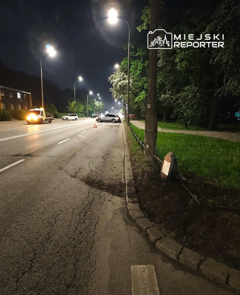 Na oświetlonej drodze nocą stoi uszkodzone auto z trójkątem ostrzegawczym, obok widać samochód pomocy drogowej.
