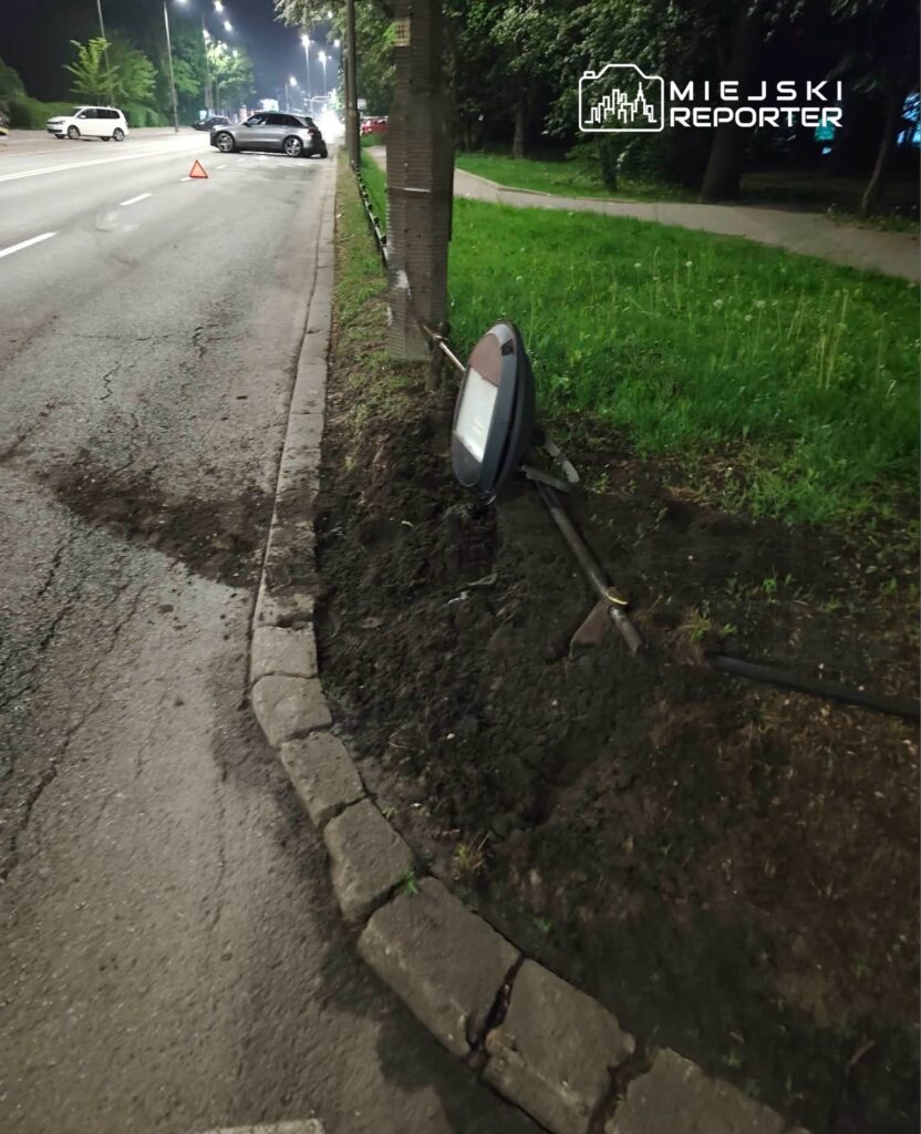 Uszkodzona latarnia uliczna leży na ziemi obok drogi, a w tle widoczny jest samochód z włączonymi światłami awaryjnymi.