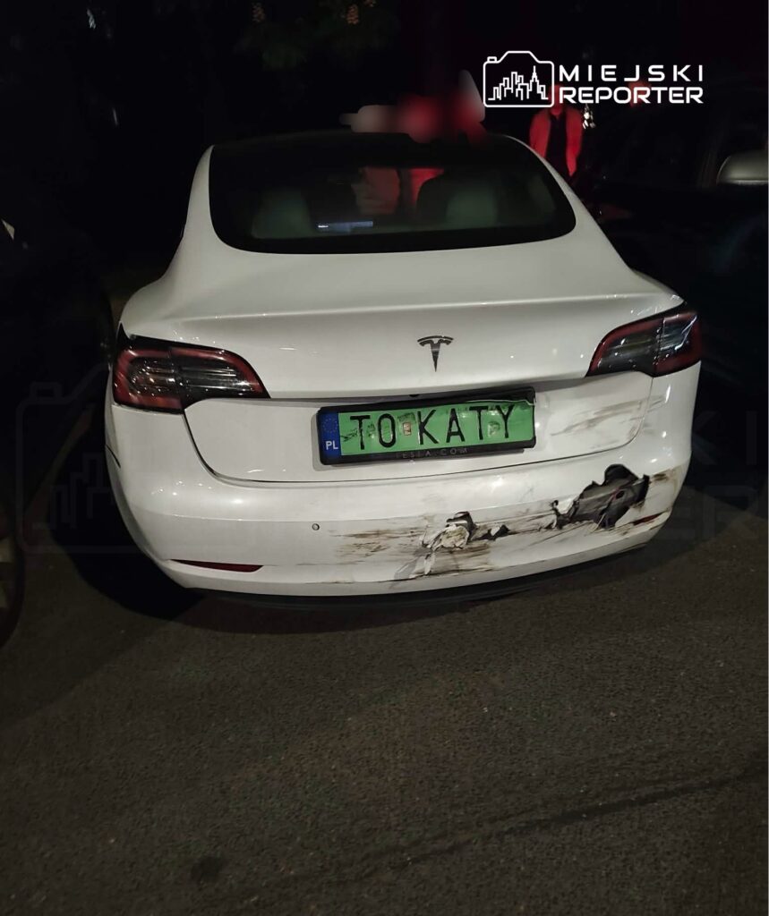 Uszkodzony biały samochód marki Tesla zaparkowany na parkingu, z widocznymi zarysowaniami na tylnej części nadwozia.