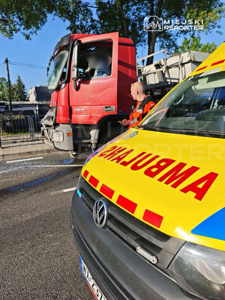 Czerwony ciężarówka stoi na drodze, obok niej mężczyzna w odblaskowym stroju rozmawia z kierowcą. W tle widać ambulanse.