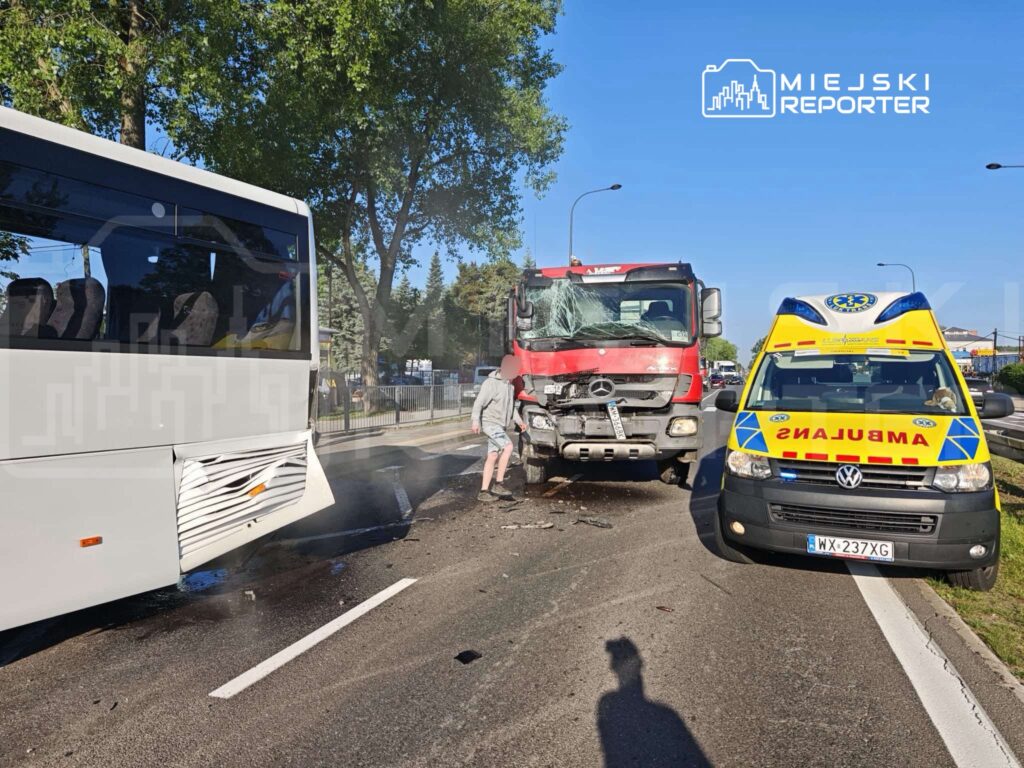 Wypadek drogowy z udziałem białego autobusu i czerwonej ciężarówki na ulicy, z widoczną ambulancją.