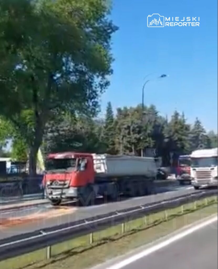 Czerwony ciężarówka z naczepą porusza się wzdłuż ruchliwej drogi, otoczonej drzewami i zielenią.