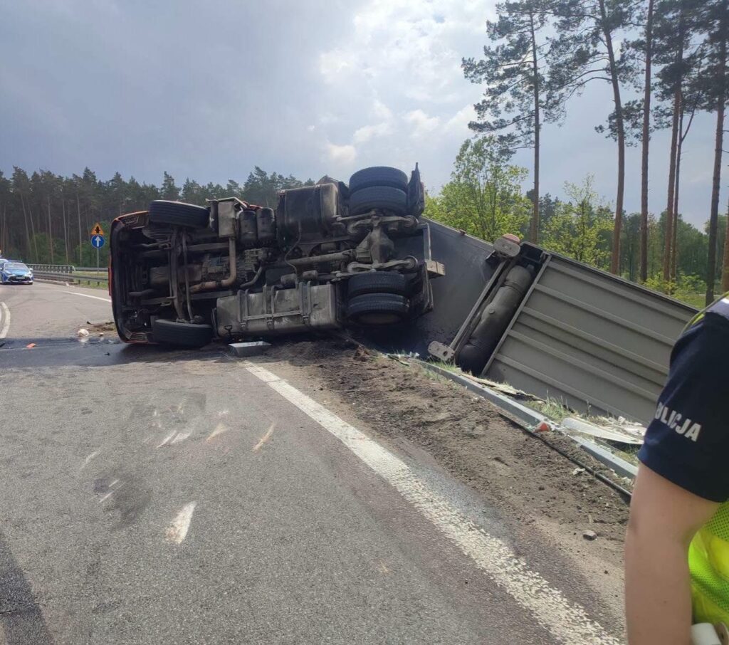 Cysterna leżąca na boku na drodze, obok stojący radiowóz policyjny i zalesiony teren w tle.