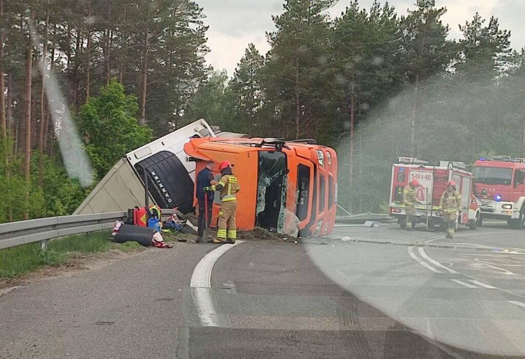 Ratownicy medyczni i strażacy pomagają przy przewróconym samochodzie ciężarowym na drodze, otoczeni przez las.