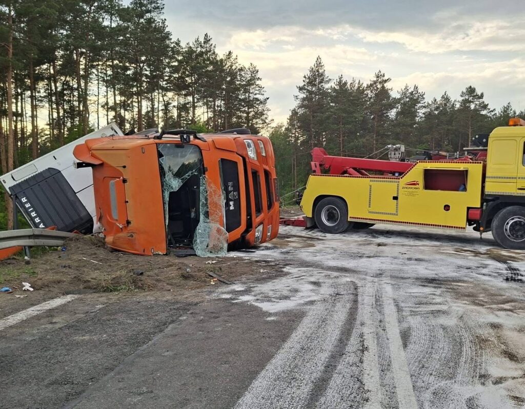 Czerwony ciężarowy pojazd leży na boku przy drodze, obok żółtej lawety, w otoczeniu lasu.