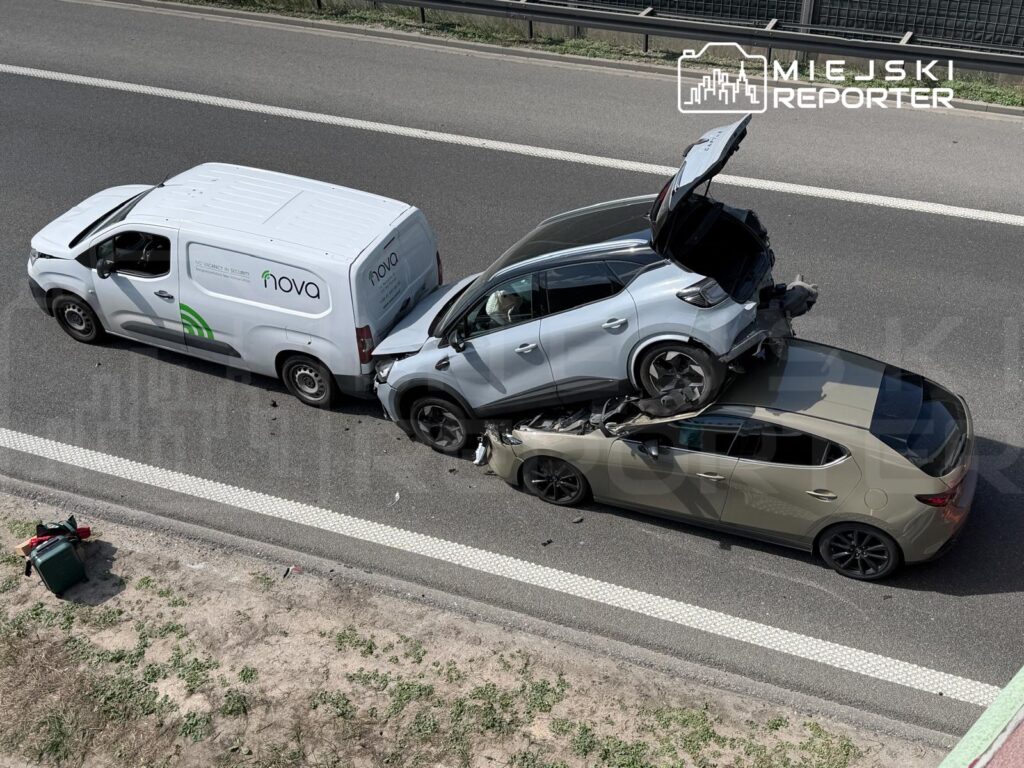 Dwa uszkodzone samochody osobowe, jeden na dachu drugiego, obok furgonetka z logo firmy, na drodze.