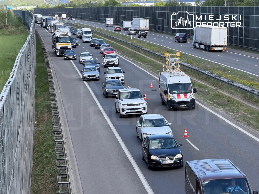 Ruch samochodowy na autostradzie z korkiem, w którym widać pojazdy osobowe oraz ciężarówki, obok furgonetka z sygnalizacją.